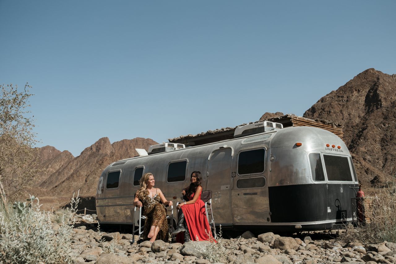 Services Two women enjoying the summer in Hatta, Dubai with a classic camper and mountainous desert backdrop.