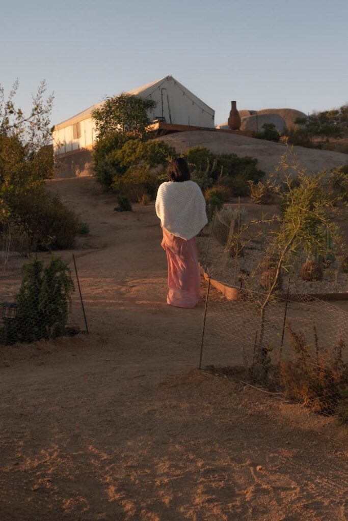 A woman wrapped in a shawl walking on a desert path towards a tent at sunrise.